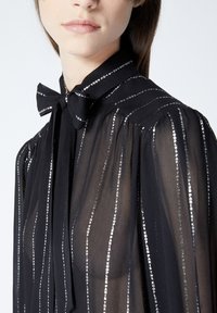 Blusa negra transparente con acentos de hilo plateado vertical, presenta un collar con lazo y mangas abullonadas, destacando una tela ligera y fluida.