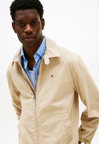 Beige zip-up jas met een omklapbare kraag, met een klein logo. Draagt over een blauw gestreept overhemd, getoond tegen een effen achtergrond.