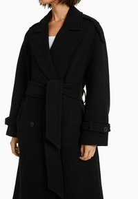 Czarny wełniany płaszcz z szerokim klapami, paskiem w talii oraz z guzikami przy mankietach o teksturowanym wykończeniu.
