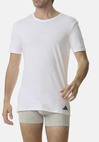 T-shirt bianco a maniche corte in cotone con colletto rotondo. Logo Adidas nero ben visibile in basso a sinistra. Modello che indossa boxer grigi.
