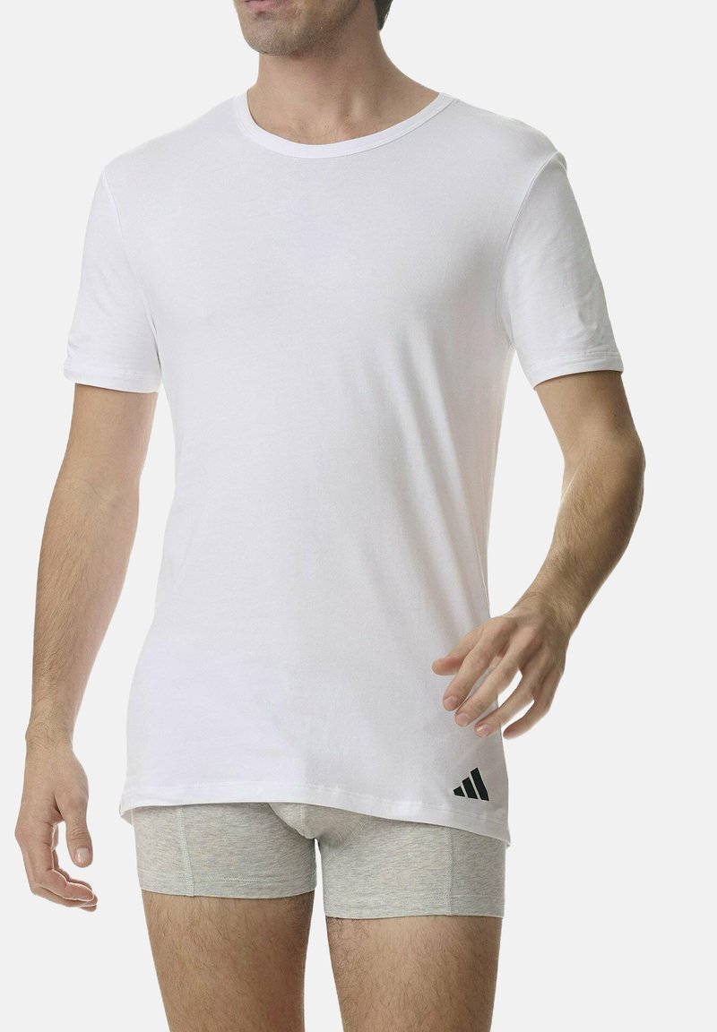 T-shirt bianco a maniche corte in cotone con colletto rotondo. Logo Adidas nero ben visibile in basso a sinistra. Modello che indossa boxer grigi.