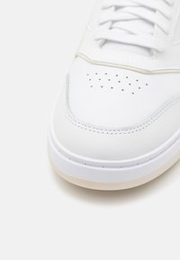 White leather sneaker with a rounded toe, perforated detailing, and textured rubber sole. Features white laces and a minimal design.