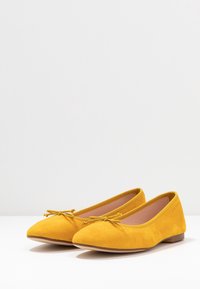 Zapatos planos de ante amarillos con punta afilada, que presentan un pequeño detalle de lazo en la parte delantera y una suela baja y lisa. El interior es de color rosa claro.