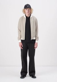 Lyse beige bomberjakke med hvid krave og manchetter, sort tekstureret polo-shirt, løstsiddende sorte bukser og sort kasket. Glat stof og afslappet pasform.