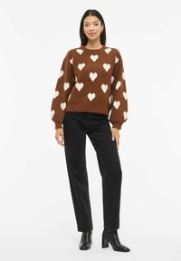 Žena stojící na bílém pozadí, oblečená v hnědém svetru s bílými srdíčky, černých rovně střižených džínách a černých kotníkových botách na podpatku.