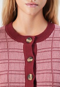 Gros plan d'une personne portant un cardigan tricoté rose et bordeaux avec trois grands boutons et un motif à carreaux.