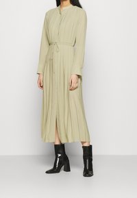 Vestito lungo plissettato in verde oliva chiaro, con collo alto, vita stretta con legaccio e maniche lunghe. Indossato con stivali neri col tacco.