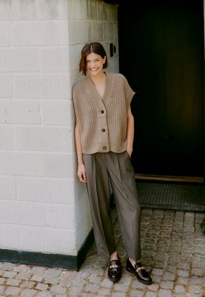 Chaleco sin mangas de punto color caqui con cuatro botones, combinado con pantalones anchos grises y mocasines negros, posando frente a una pared de piedra.