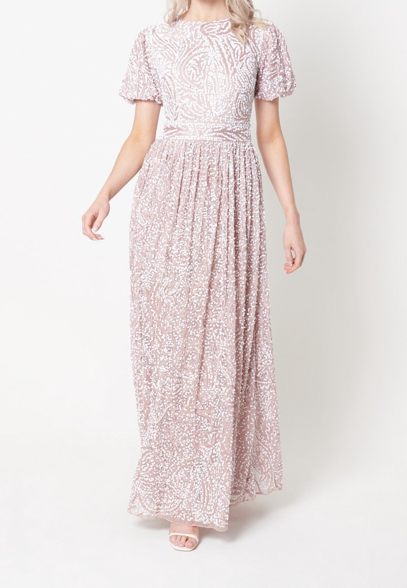 Femme portant une robe longue rose clair avec des motifs en sequins blancs et des manches courtes bouffantes, debout sur un fond blanc.