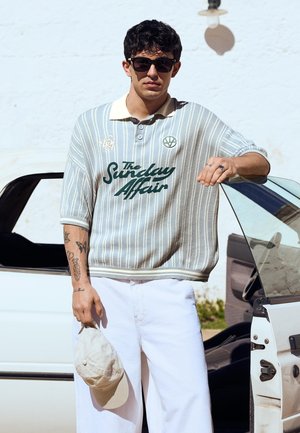 Giovane uomo che indossa occhiali da sole e una polo a righe con la scritta "The Sunday Affair", tenendo un cappellino beige, in piedi accanto a una portiera aperta dell'auto.