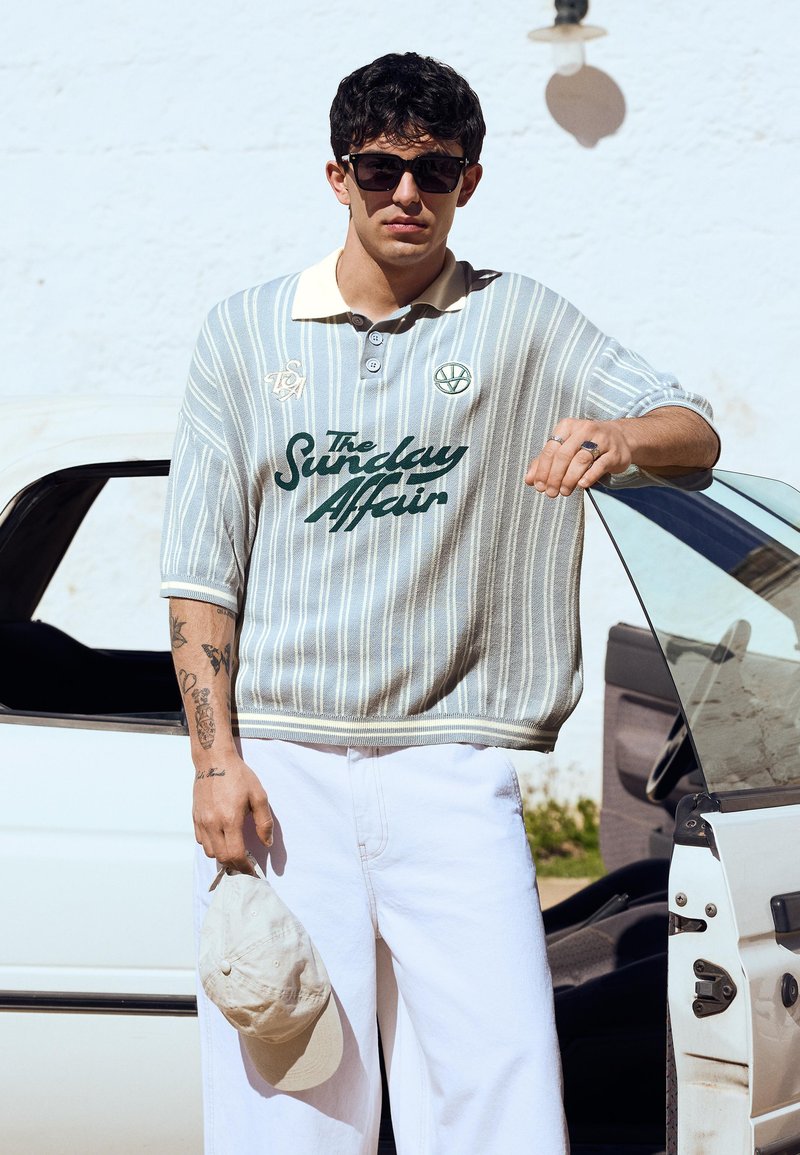 Giovane uomo che indossa occhiali da sole e una polo a righe con la scritta "The Sunday Affair", tenendo un cappellino beige, in piedi accanto a una portiera aperta dell'auto.