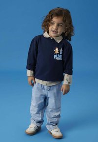 Marineblå hettegenser med rullede ermer har tegneseriegrafikk. Lyse denimbukser med elastisk midje. Hvite sneakers med brune såler.