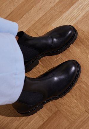 Czarne skórzane botki za kostkę z elastycznymi panelami bocznymi, teksturowaną gumową podeszwą i błyszczącym wykończeniem, umieszczone na drewnianej podłodze.
