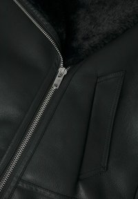 Veste en cuir noire avec fermeture éclair argentée partiellement ouverte, montrant une doublure en fourrure douce noire et une poche cousue à l'avant.