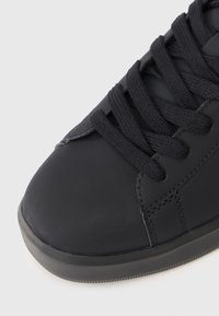 Gros plan sur une sneaker noire avec un bout arrondi, des lacets en tissu noir et un dessus en cuir cousu sur fond blanc.
