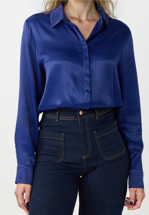 Vrouw draagt een lange mouwen koningsblauwe satijnen blouse, ingestopt in een hoog tailleren donkerblauwe spijkerbroek met grote voorzakken en contraststiksels.