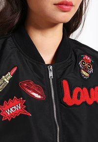 Czarna kurtka bomber z kolorowymi, haftowanymi naszywkami, w tym pomadką, ustami i czaszką, ze srebrnym zamkiem błyskawicznym i ściągaczem na kołnierzu.