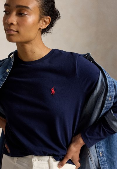 Γυναίκα που φοράει μπλε σκούρο πουκάμισο με μακριά μανίκια με κόκκινο κεντημένο λογότυπο αλόγου, λευκό παντελόνι και μερικώς φορεμένο τζιν μπουφάν.