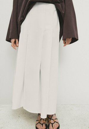 Lower body wearing a long white skirt with front pleat, brown long-sleeve top, and black strappy sandals standing on speckled floor.