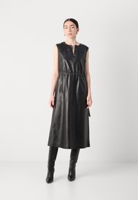 Schwarzes Lederkleid ohne Ärmel mit V-Ausschnitt, geraffter Taille und lang fließender Silhouette. Kombiniert mit knielangen schwarzen Stiefeln.