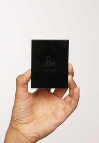 Svart firkantet kompakt med matt overflate, med preget "dm" og "Danessa Myricks Beauty" logoer på forsiden. Holdes i en hånd.
