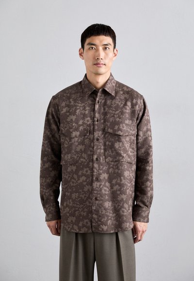 Camicia marrone con motivo, dotata di due tasche frontali, colletto button-down e maniche lunghe, realizzata in tessuto texture, abbinata a pantaloni grigi chiari.