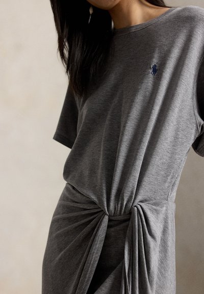 Mujer con vestido gris de manga corta y detalle fruncido con nudo en la cintura, con un pequeño logo bordado en el pecho.