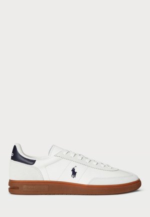 Zapatilla blanca con suela de goma marrón, parte superior de textil, acento en el talón en azul marino y detalle de logo en el lado. Cuenta con seis ojetes y cordones planos.