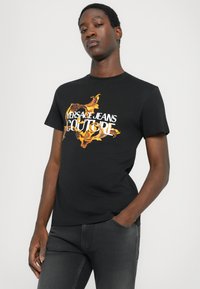 Črna bombažna majica s kratkimi rokavi in belim ter oranžnim grafičnim tiskom "Versace Jeans Couture". Ima krožen izrez in prilegajočo se zasnovo.