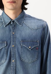 Camicia di denim azzurro chiaro con colletto punta, due tasche sul petto con bottoni e chiusure a scatto metalliche, caratterizzata da una trama sottile e un dettaglio del logo rosso.
