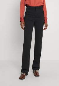 Zwarte op maat gemaakte broek met een recht model, gemaakt van een gladde stof. Bevat een platte voorkant en riemlussen. Gepaarde met bruine hakken.