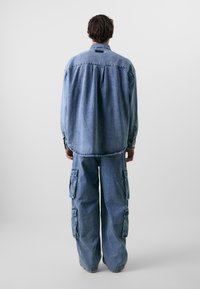 Camisa de denim e calças cargo largas em azul claro. A camisa tem punhos com botão e yoke nas costas; as calças têm bolsos laterais e um ajuste relaxado.