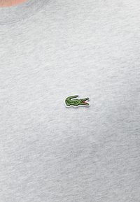 Gråt stof med en lille grøn broderet krokodille-logo med åben mund, syet på brystområdet af et stykke tøj.