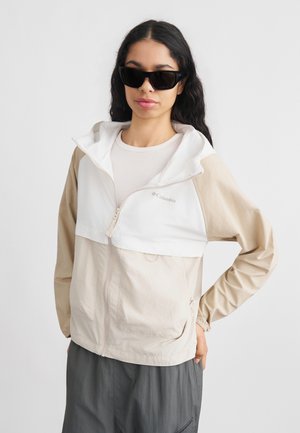 Kvinde med langt mørkt hår iført sorte solbriller, beige og hvid Columbia-jakke samt grå bukser, stående med den ene hånd på hoften.