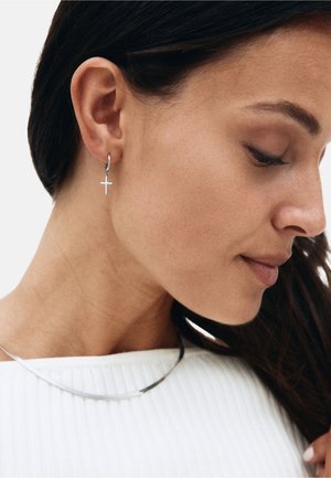 Zilveren creolen met een klein kruis detail. De drager heeft een egale huid en bruin haar, draagt een witte ribbel top en een zilveren ketting.