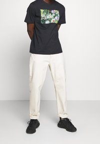 Černé bavlněné tričko s barevným květinovým potiskem na přední straně. Spárováno se smetanovými kalhotami a černými teniskami.