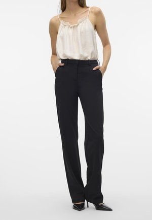 Femme avec les mains dans les poches d'un pantalon noir, portant un haut blanc sans manches à volants et des talons hauts noirs à bout pointu.