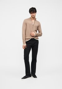 Pull beige à col zippé avec manches longues, associé à un pantalon noir à jambes droites et des baskets noires. Détail logo subtil sur le pull.