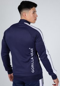 Marine sportjack met hoge kraag, witte zijstrepen en logo op de achterkant. Glad oppervlak, met ritszak, aansluitend ontwerp.