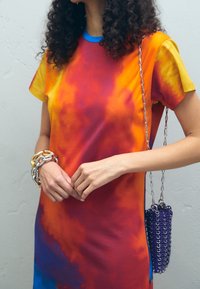 Barevné tričkové šaty s efektom tie-dye v žlutých, oranžových a červených odstínech, s krátkými rukávy a modrým výstřihem; v kombinaci se stříbrným řetízkovým náramkem a fialovou kabelkou.