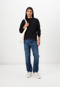 Black sweatshirt with ribbed cuffs, worn over a navy shirt. Paired with blue denim jeans and white sneakers. Holding a beige tote bag.