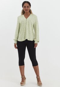 Lichtgroen blouse met een V-hals, lange mouwen en een gestructureerd patroon van kleine bloemetjes; gecombineerd met zwarte cropped leggings en sleehakken.