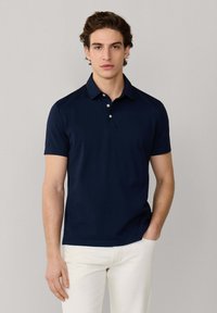 Joven vestido con una camisa polo de manga corta azul marino y pantalones blancos, de pie con una mano en el bolsillo contra un fondo neutro.