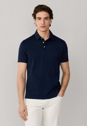 Joven vestido con una camisa polo de manga corta azul marino y pantalones blancos, de pie con una mano en el bolsillo contra un fondo neutro.