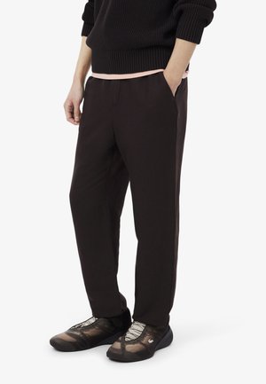 Persoon draagt zwarte broek, zwarte trui met roze zoom zichtbaar, en zwarte mesh sneakers met witte veters, staat met één hand in de zak.