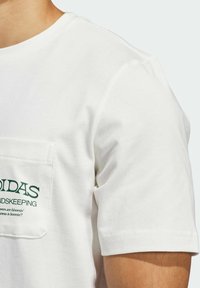 Bílý bavlněný tričko s kulatým výstřihem, krátkými rukávy a náprsní kapsou s zeleným textem a logem. Hladká textura, ležérní design.