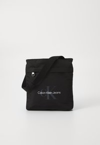 Borsa a spalla in nylon nero con tracolla regolabile, caratterizzata dal logo "Calvin Klein Jeans" in bianco e grigio sulla parte frontale.
