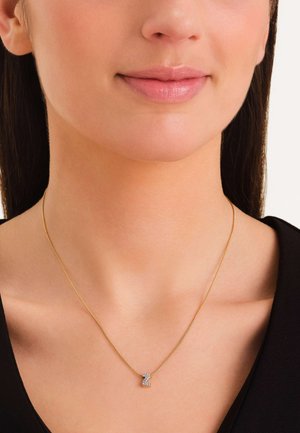 Gouden ketting met een verfijnde schakel en een met rhinestones versierde letterhanger in cursieve stijl, dicht bij het sleutelbeen gedragen.