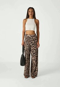 Kvinna står upprätt, klädd i vit ärmlös croptop, bredvid byxor med leopardsmönster, svarta sandaler och håller i en svart handväska.