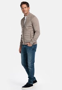 Man staat met hand in zak, draagt een beige gewatteerde ritsjack, blauwe jeans en zwarte sneakers tegen een witte achtergrond.